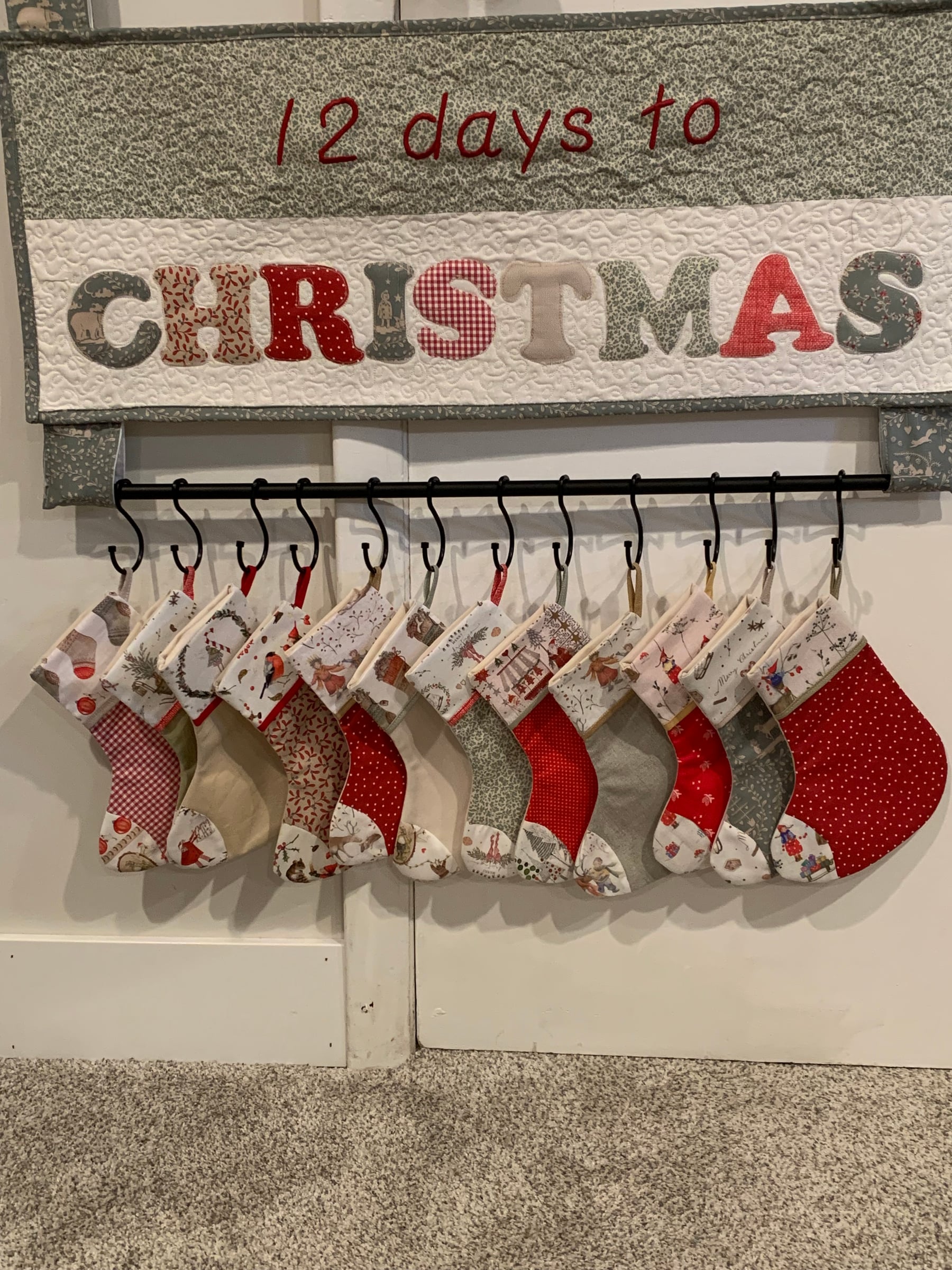 The 12 Days to Christmas banner on a white wall, with twelve mini stockings hanging from a black rod beneath it